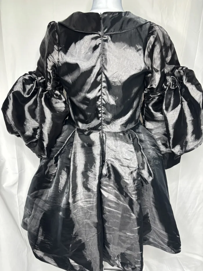 The Women Metallic Organza Black Pleated Dress features voluminous puffed long sleeves, a gathered flared skirt, and a back zipper, shown on a mannequin against a plain white background.