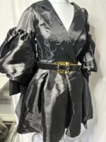 The Women Metallic Organza Black Pleated Dress features shiny black fabric, long puffed sleeves, a wide notched collar, a pleated skirt, and a thick black belt with a large gold buckle, displayed on a mannequin.