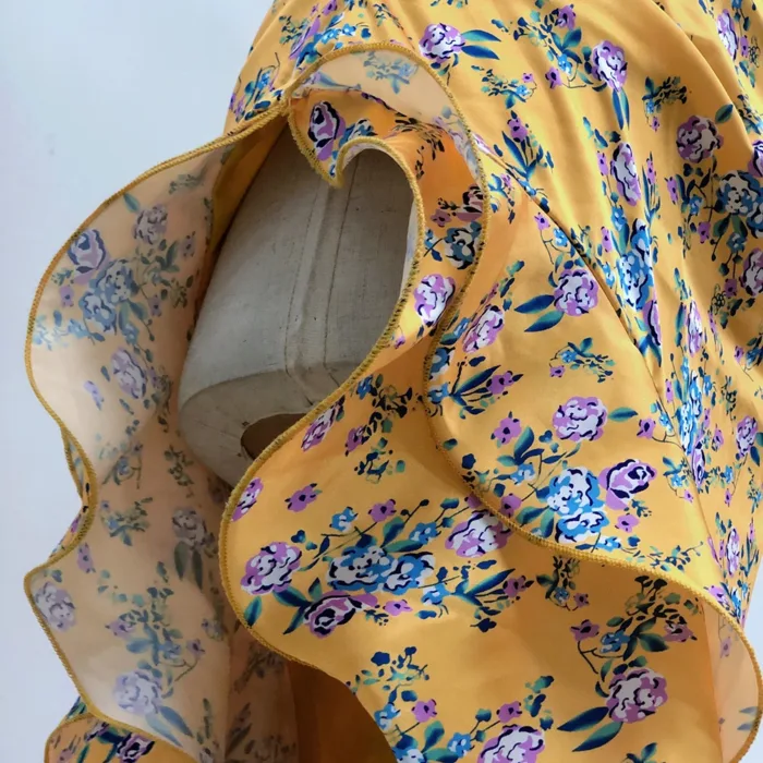 A close-up of a Hollowed-Out Hip Irregular Ruffled Floral Dress displayed on a mannequin, featuring yellow fabric with blue, purple, and white flowers and wavy ruffled edges.