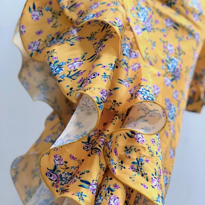 A close-up of the Hollowed-Out Hip Irregular Ruffled Floral Dress shows yellow fabric with blue and purple flowers, highlighted by wavy, ruffled edges for a flowing, dynamic look.