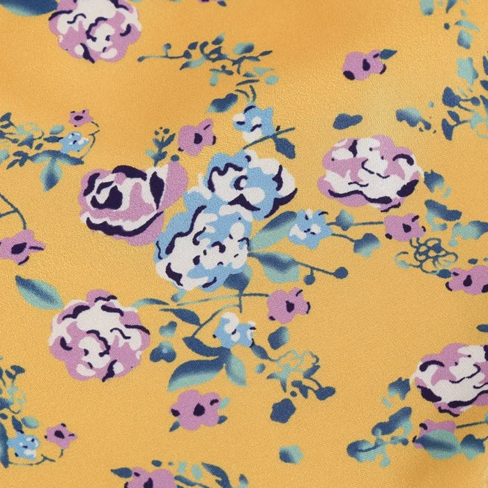 A close-up of the Hollowed-Out Hip Irregular Ruffled Floral Dress shows yellow fabric with pink, white, and blue flowers and green leaves scattered across the surface.
