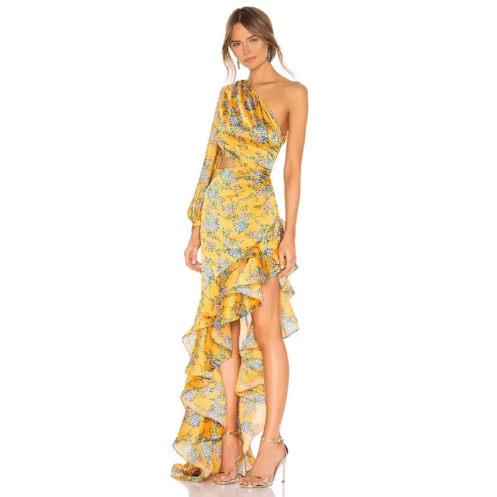 A woman stands against a white background wearing the Hollowed-Out Hip Irregular Ruffled Floral Dress with yellow, blue floral patterns, an asymmetrical one-shoulder design, ruffled high-low hem, and silver high-heeled sandals.