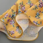 A close-up of the Hollowed-Out Hip Irregular Ruffled Floral Dress’s yellow fabric, featuring a purple, blue, and green floral pattern and neatly finished yellow stitching along the edge, with both front and back sides visible.