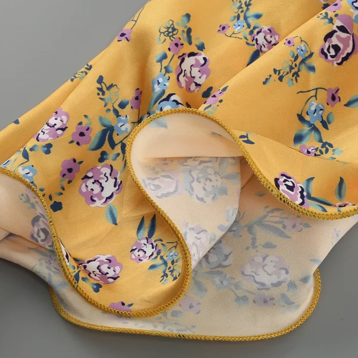 A close-up of the Hollowed-Out Hip Irregular Ruffled Floral Dress’s yellow fabric, featuring a purple, blue, and green floral pattern and neatly finished yellow stitching along the edge, with both front and back sides visible.