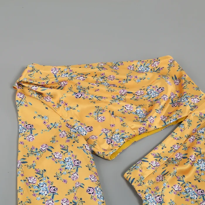 The Hollowed-Out Hip Irregular Ruffled Floral Dress is displayed flat on a gray surface, showcasing its yellow fabric adorned with small pink, blue, and white flowers featuring green leaves.
